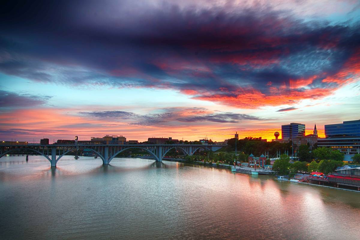 Vivid sunset over the Tennessee River at Knoxville, with an arched bridge silhouetted against orange and red cloud and the downtown skyline beyond.